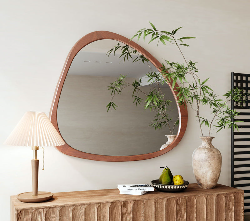 Asymmetrical wood wall mirror over a sideboard with a table lamp and greenery