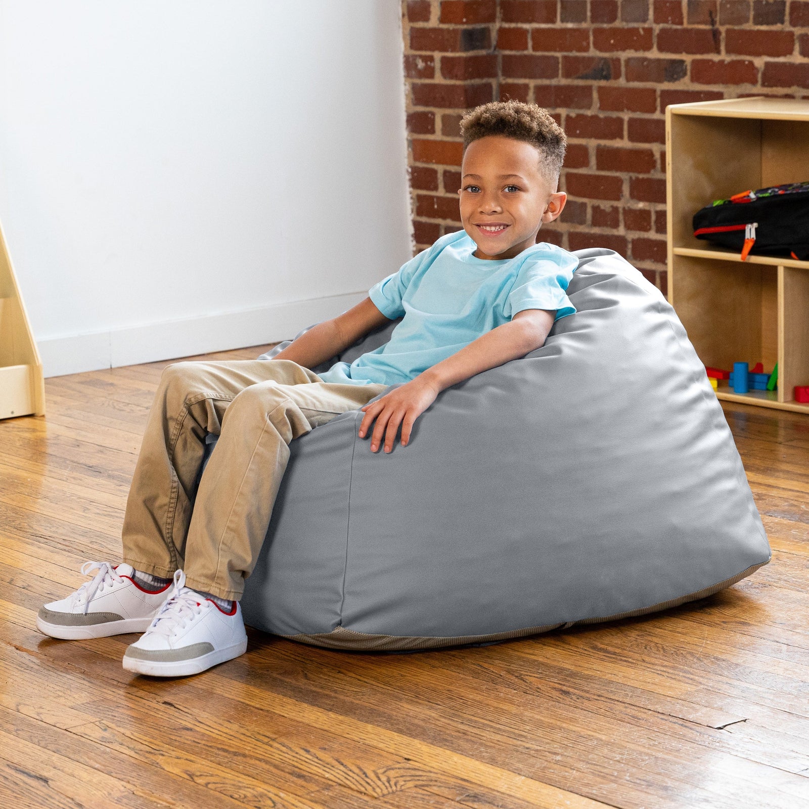 Child sitting on a gray bean bag chair in a room with a brick wall and wooden floor.