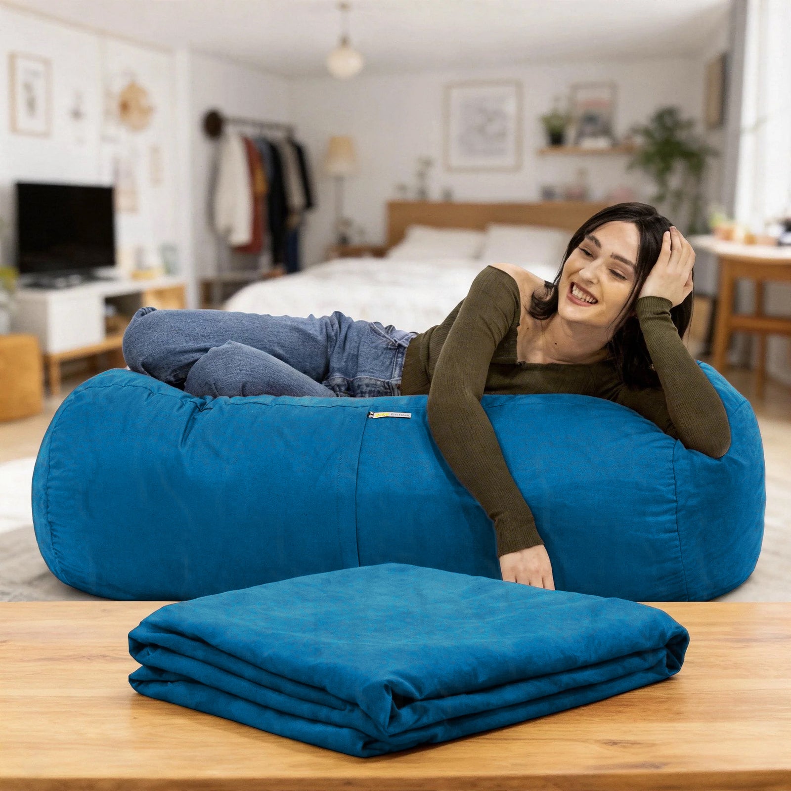 A woman relaxes on a large teal-blue bean bag chair in a bright bedroom, with a matching folded blanket placed on a wooden table in the foreground.