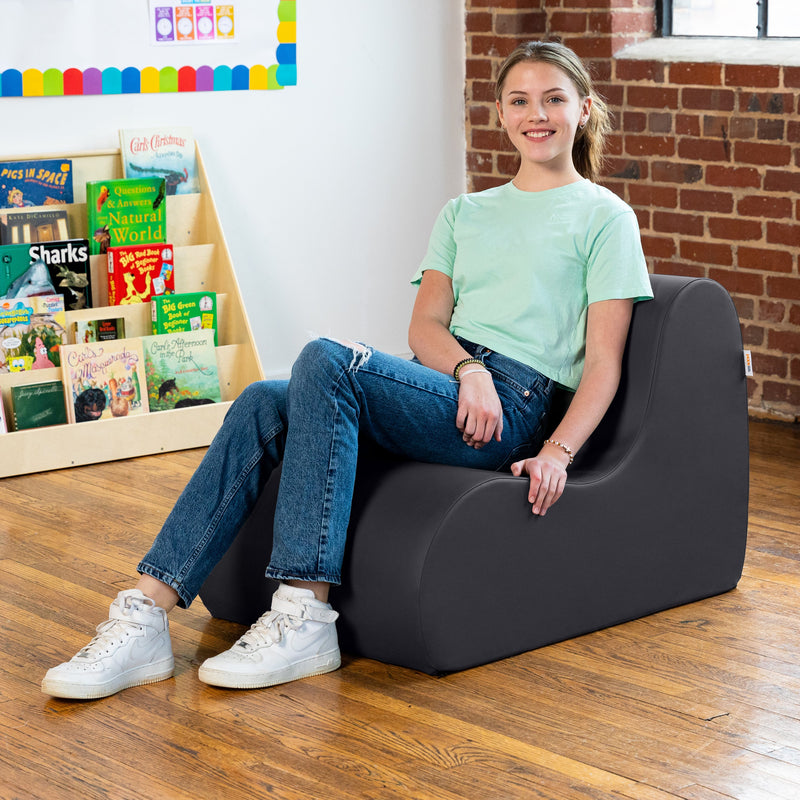 Young girl sitting on a gray chair in a room with books and a brick wall.