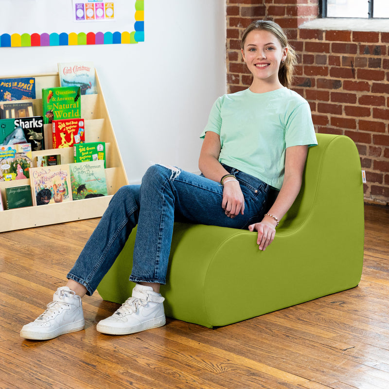 Young girl sitting on a green chair in a room with books and a brick wall.