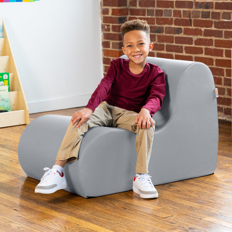 Child sitting on a gray children's chair in a room with a brick wall and wooden floor.