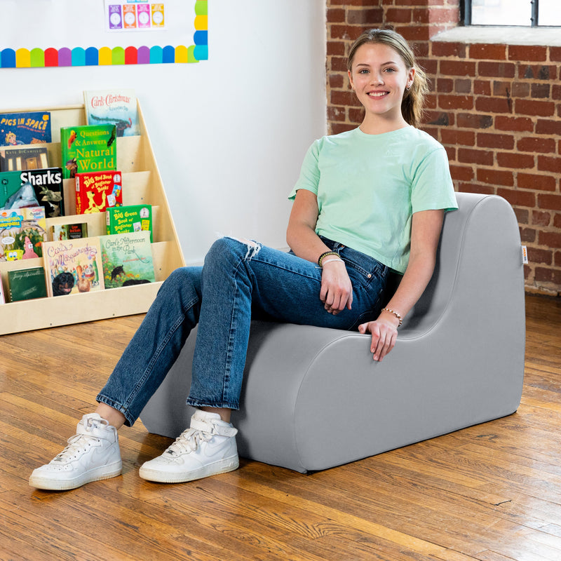 Young girl sitting on a gray chair in a room with books and a brick wall.