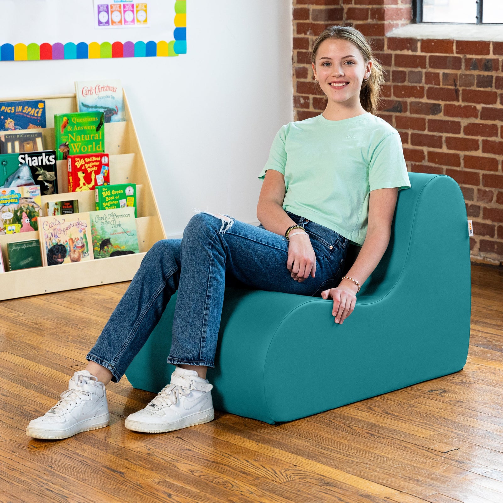 Young girl sitting on a teal chair in a room with books and a brick wall.